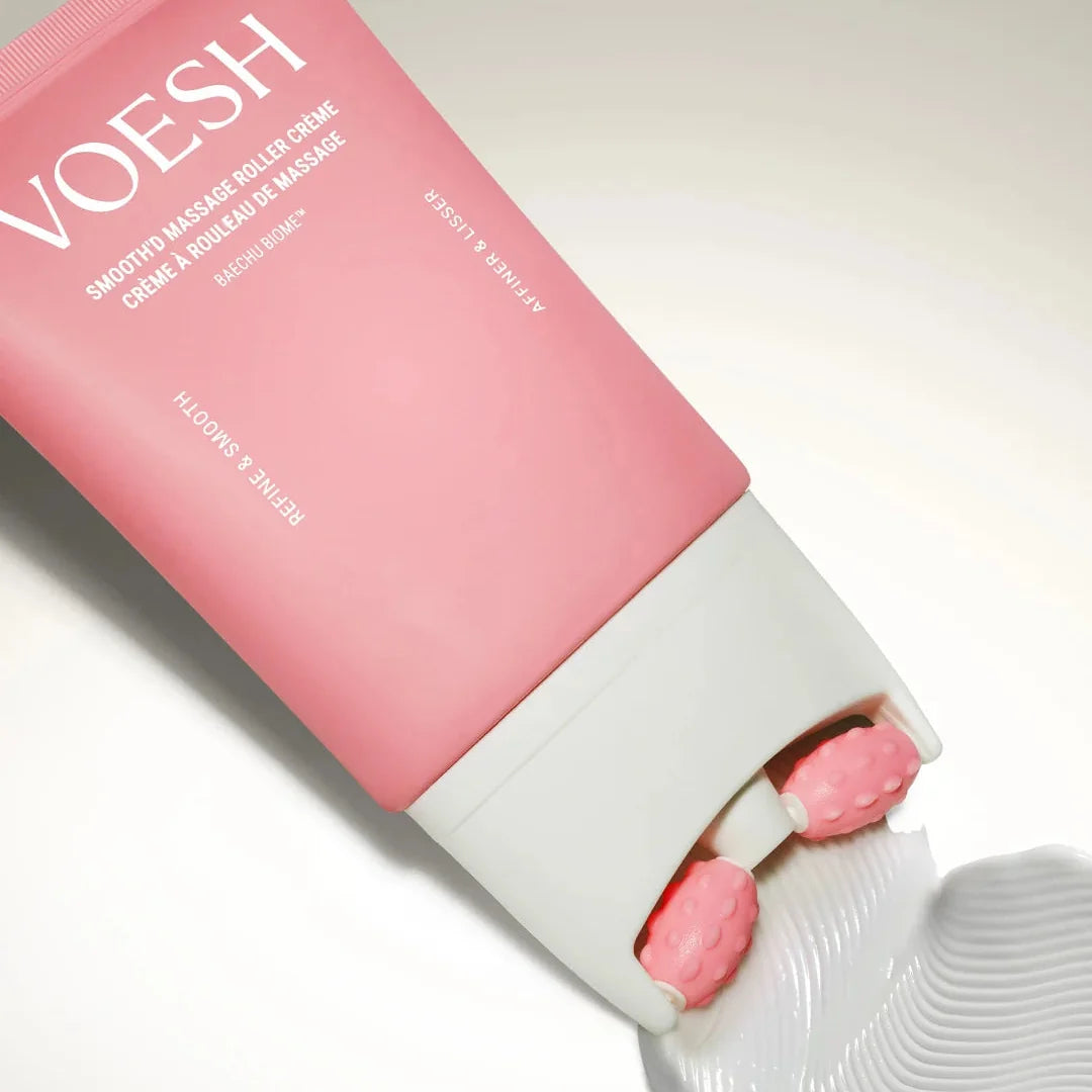 A pink tube of VOESH Smoothed Brazilian Melinica Creme is shown diagonally with the white cap unscrewed, releasing two pink gel beads onto a white surface that has a textured smear of the white cream.
