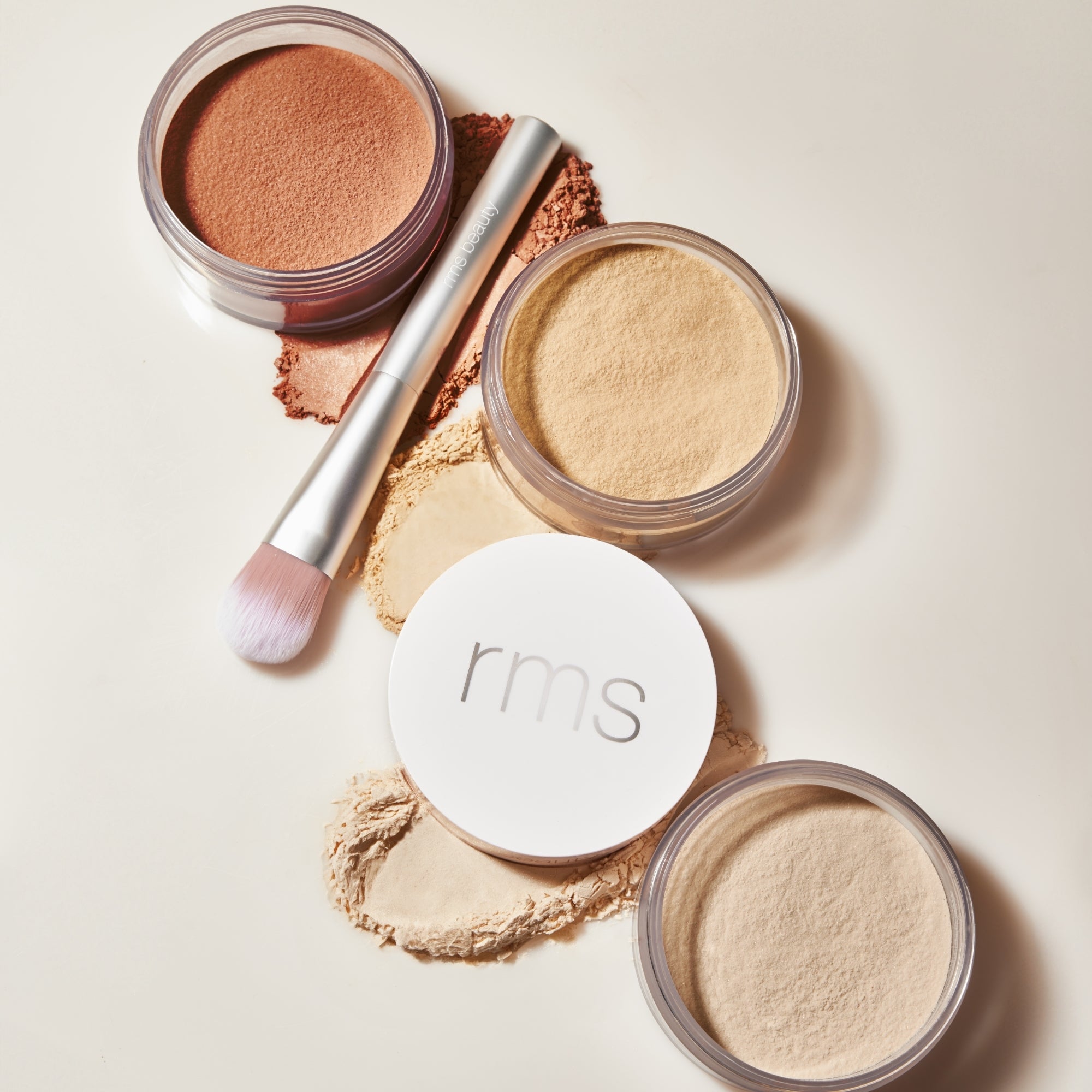 Four jars of RMS beauty powder with a brush on a light background