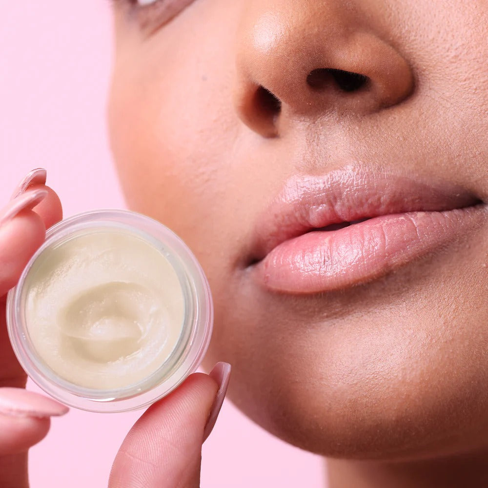 Close-up of a person holding a small container of NCLA Lip Balm near their face on a pink background.