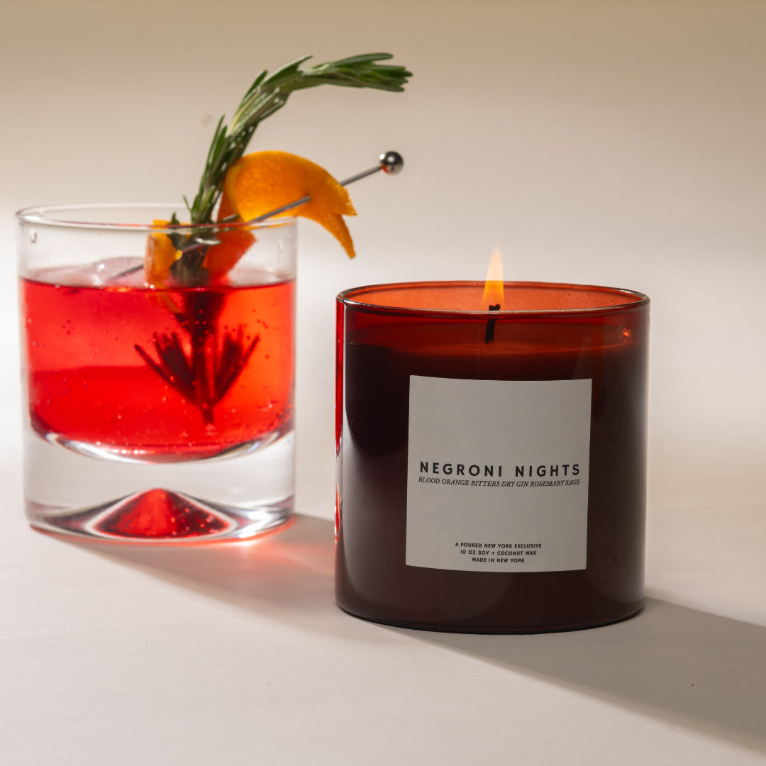 Candle labeled 'Negroni Nights' next to a red cocktail with an orange and rosemary garnish.
