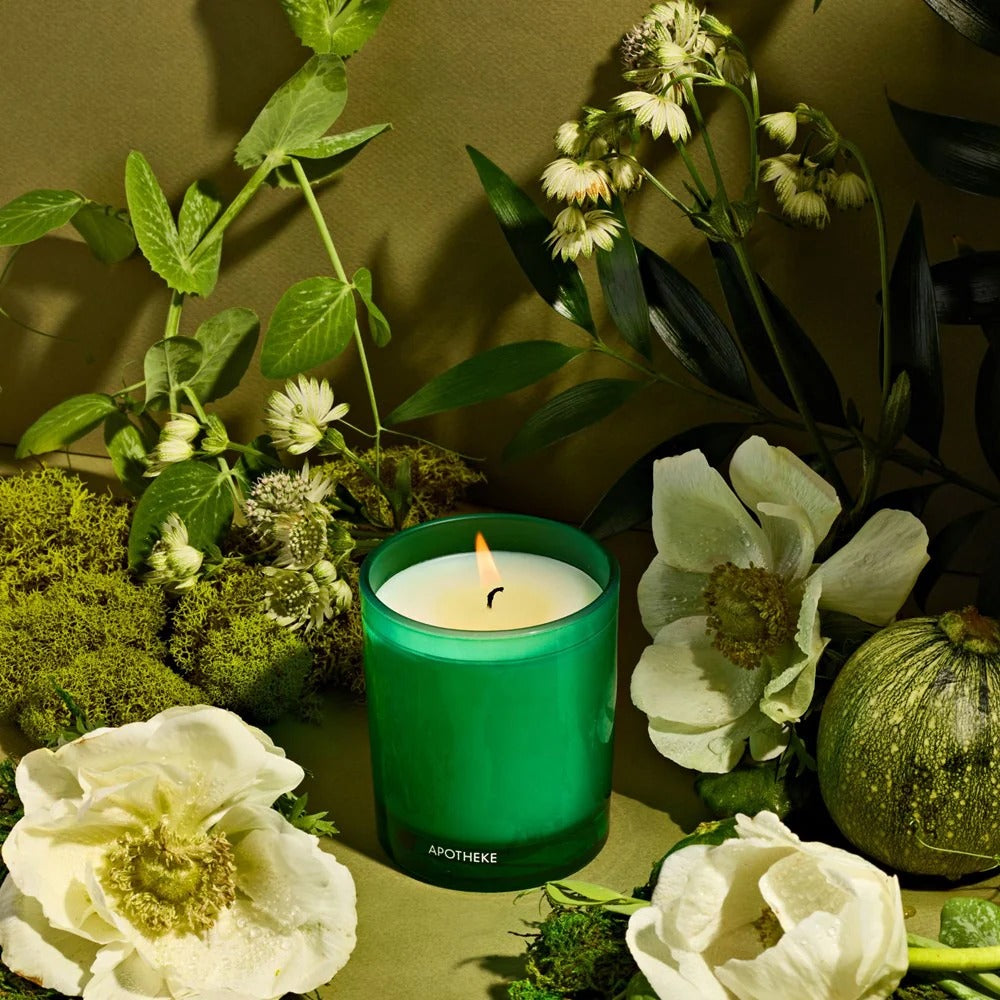 Verdant Garden Petite Candle in a green vessel on a green background surrounded by herbs, flowers, botanicals, mosses and melons. 