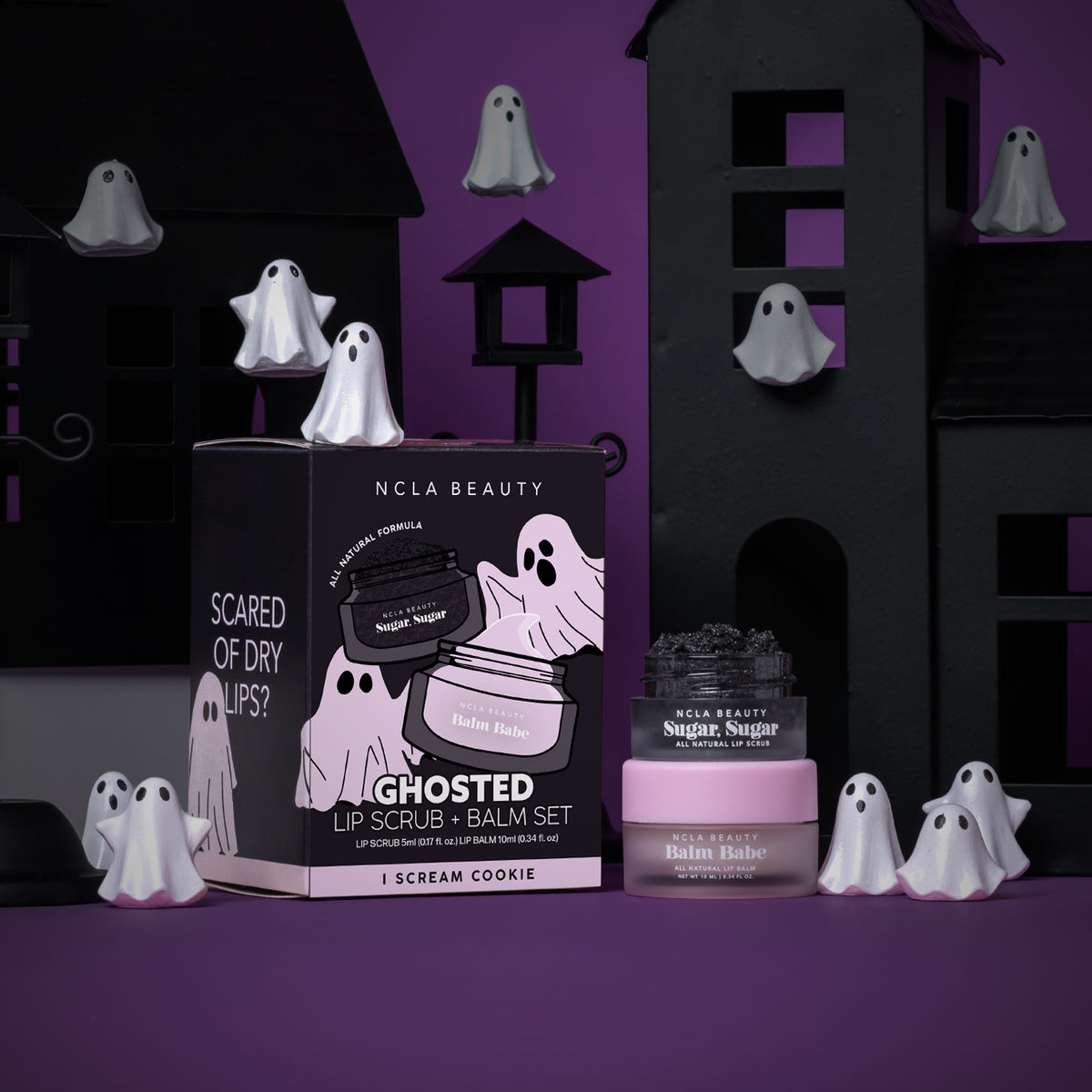 NCLA Beauty Ghosted Lip Scrub and Balm Set in 'I Scream Cookie' flavor, displayed with its black and pink packaging box and the two jars (one with a pink lid, one with a black lid) against a Halloween-themed background of black haunted houses and white ghost decorations on a purple surface.