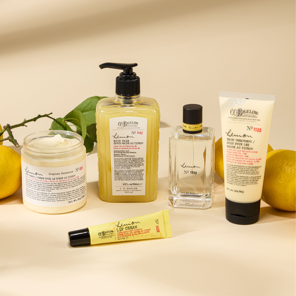 Collection of Bigelow's skincare products including a jar, bottle, lotion, and lip balm on a beige background with lemons and leaves.