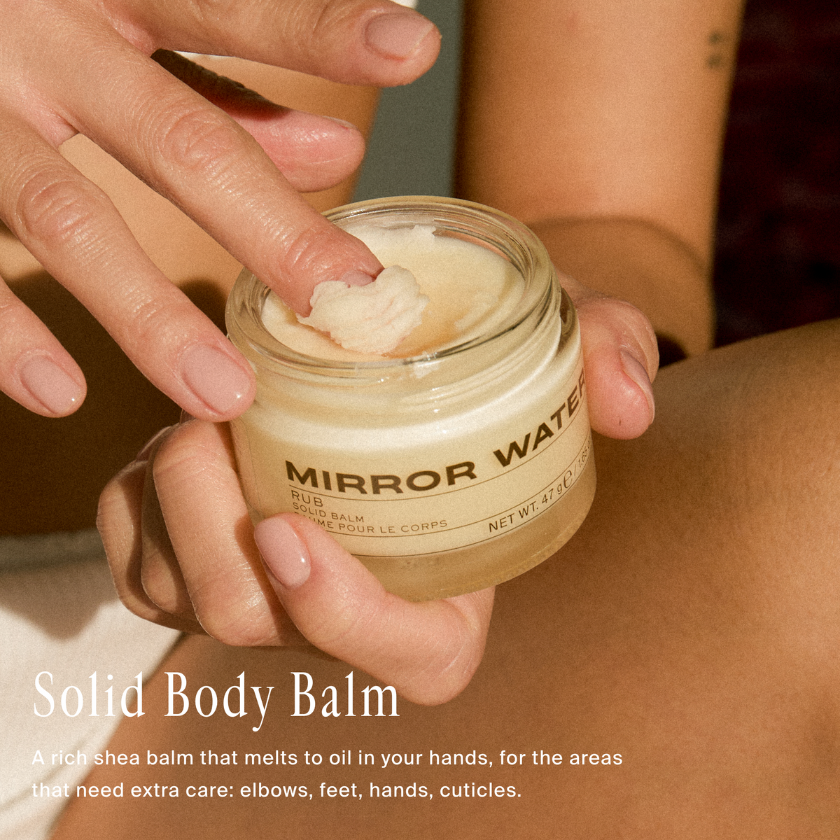 A close-up of a person holding a small clear jar of Mirror Water Solid Body Balm, with their fingers touching the cream inside. The jar has a beige label with black text.