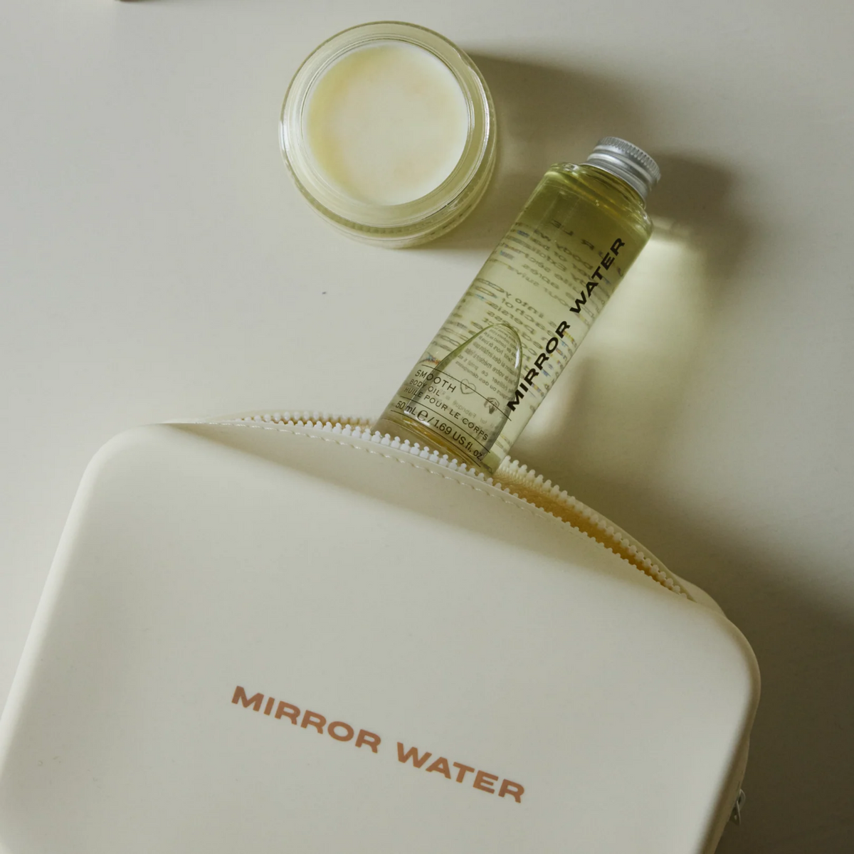A cosmetic travel set featuring a cream-colored zippered pouch with 'MIRROR WATER' printed in orange, a clear bottle of Mirror Water serum with a silver cap, and an open jar of white cream, arranged on a light beige surface.