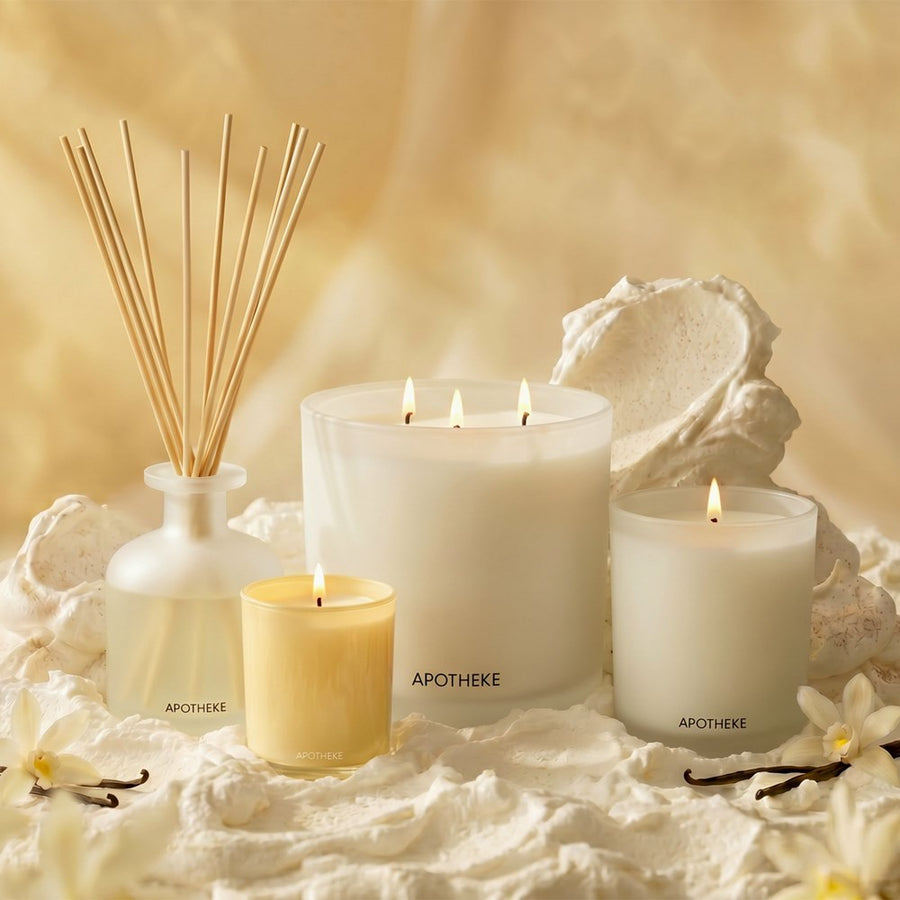 A styled arrangement of Apotheke home fragrance products on a soft cream surface, including a reed diffuser bottle with wooden reeds, a large white candle, a smaller yellow candle, and a white candle jar, with decorative shells and vanilla beans.