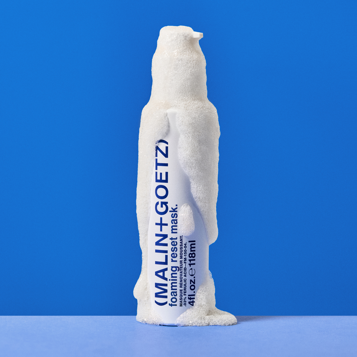 Bottle of Malin+Goetz Foaming Reset Mask covered in foam on a light blue surface in front of a darker blue background