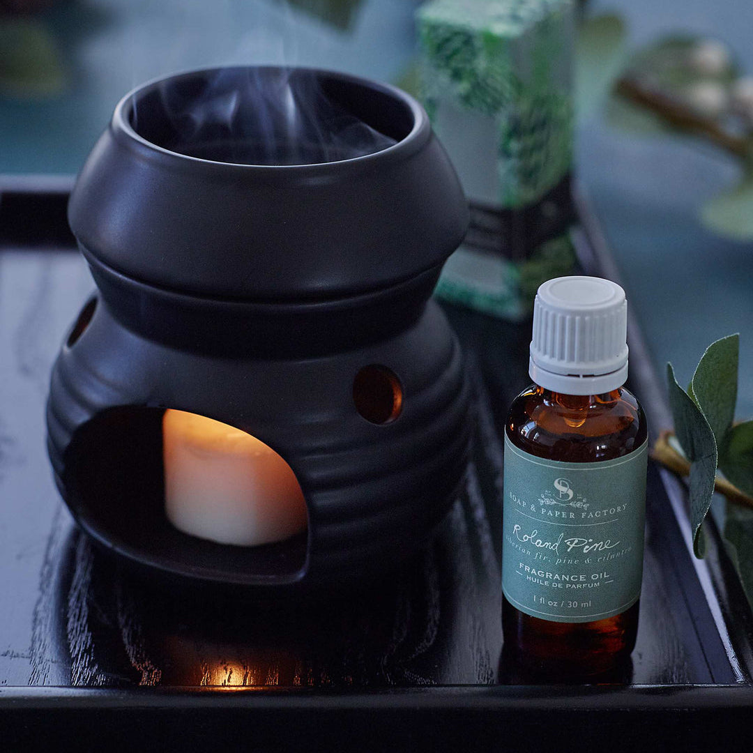 Black ceramic diffuser with a lit candle and a bottle of Roland Pine fragrance oil on a dark surface.