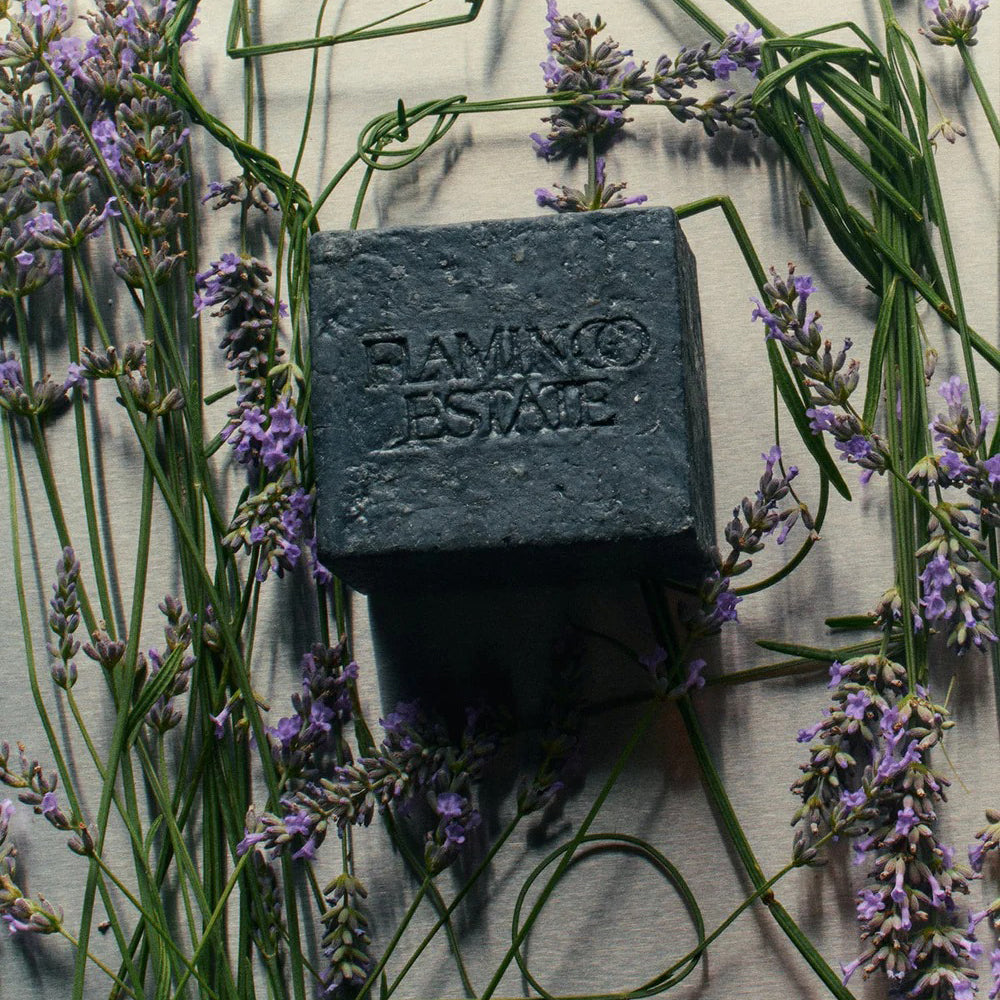 Charcoal Bar of soap with 'Flamingo Estate ' branding surrounded by lavender flowers on a textured surface.