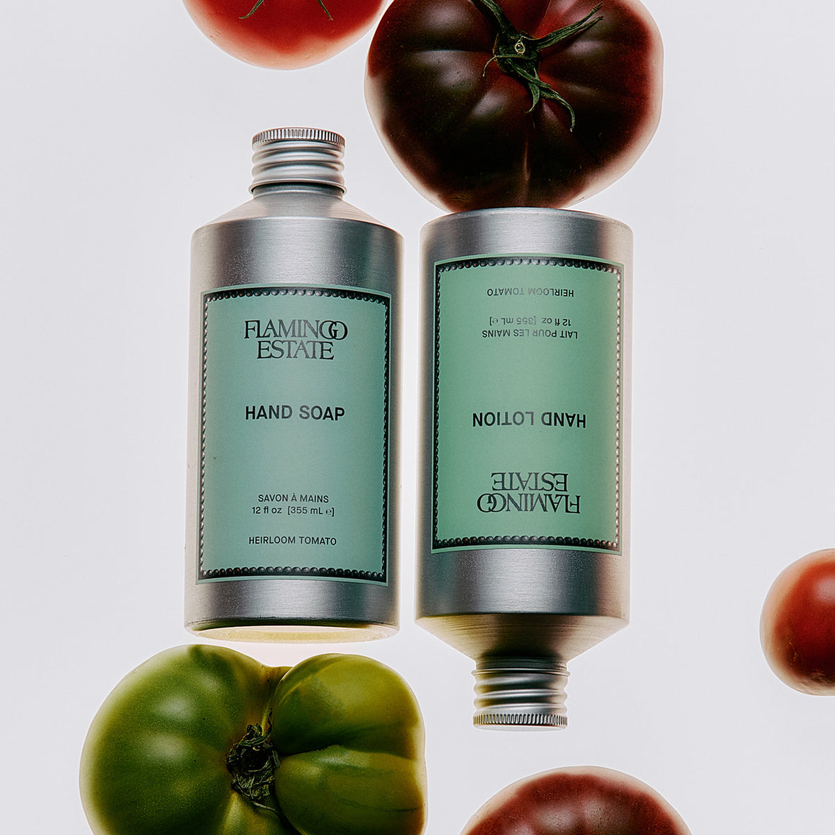 Two silver tubes of Flamingo Estate hand care products, one labeled Hand Soap and one labeled Hand Lotion, with mint green labels, arranged around whole tomatoes on a light gray background.