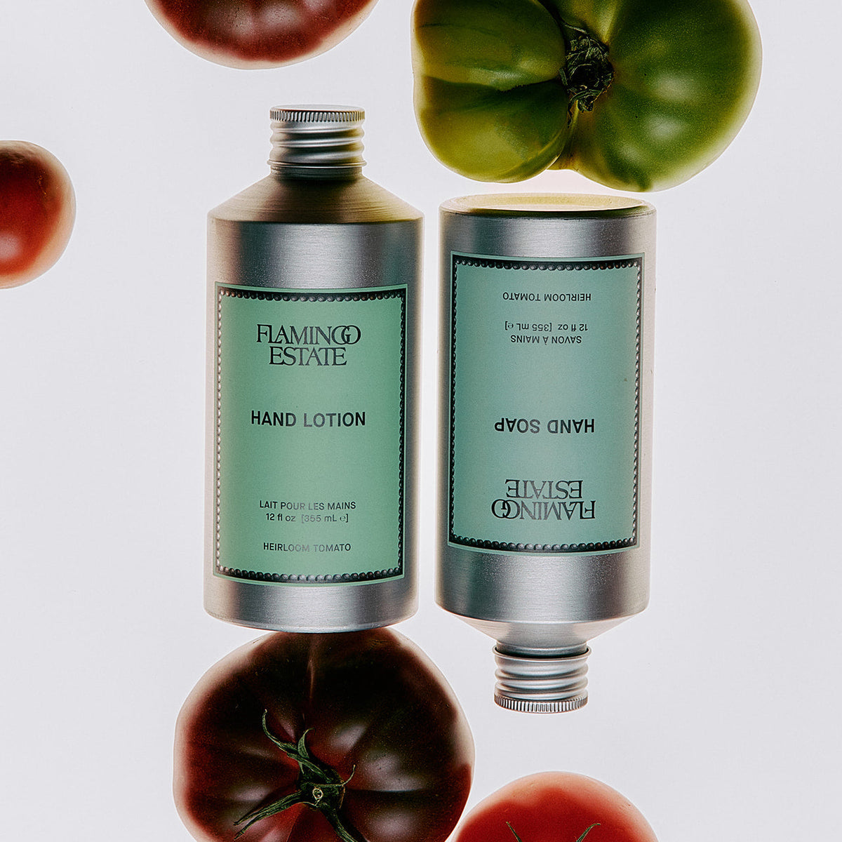 Two silver tubes of Flamingo Estate hand care products, one labeled Hand Soap and one labeled Hand Lotion, with mint green labels, arranged around whole tomatoes on a light gray background.