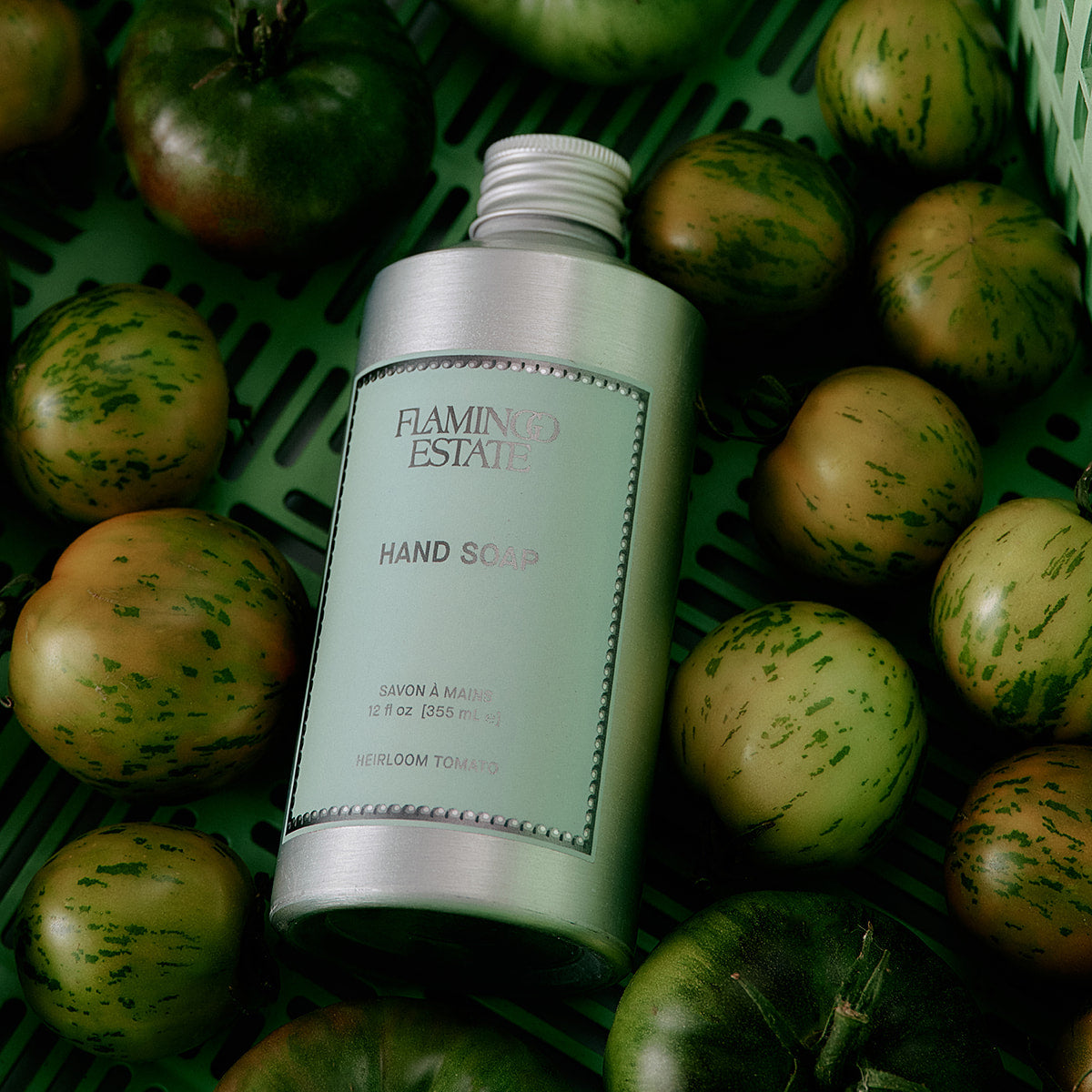 Flamingo Estate hand soap bottle surrounded by green tomatoes on a green surface