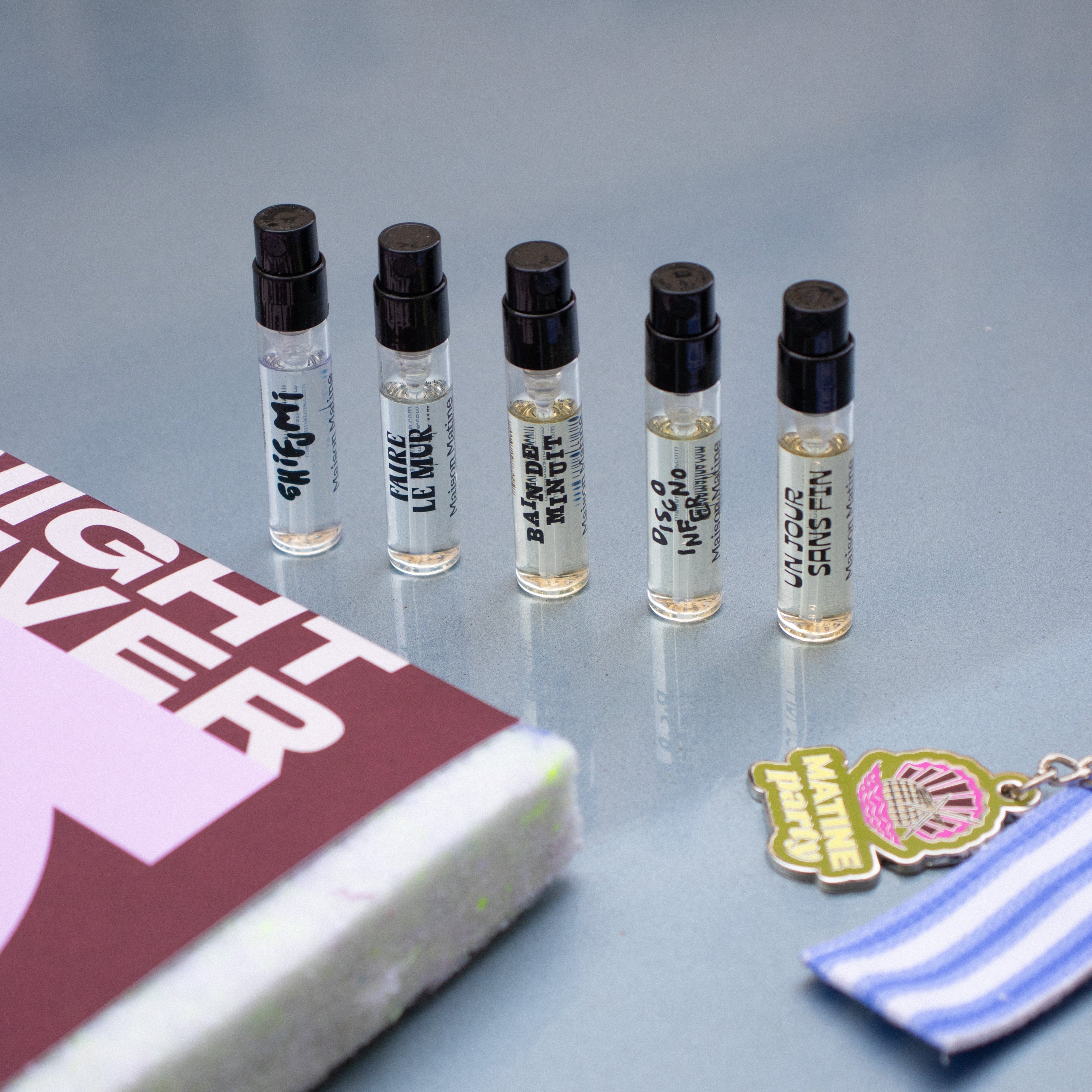 Five small perfume bottles with black caps on a gray surface, next to a book and a keychain.