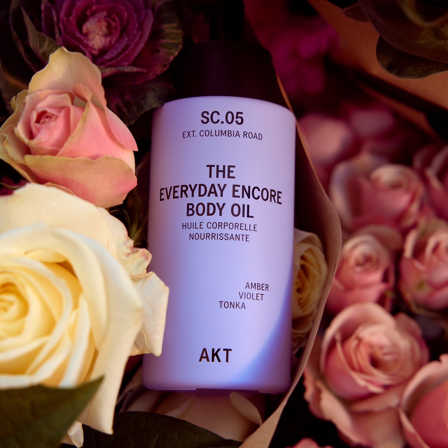 A bottle of AKT The Everyday Encore Body Oil in scent SC.05 Amber Violet Tonka, styled in a flat-lay arrangement with pink and cream roses.