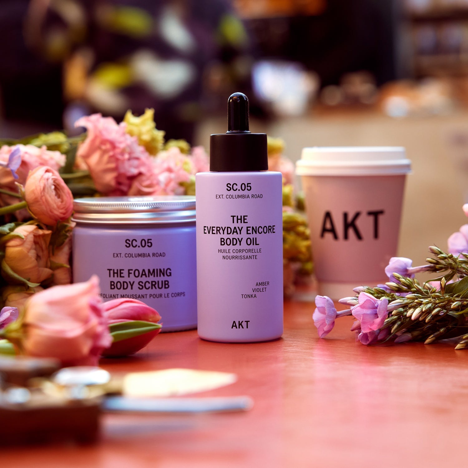 The AKT Everyday Encore Body Oil Sc.05 including a lavender body scrub in a tin, and a pink coffee cup with AKT branding, styled with pink roses on a wooden table.