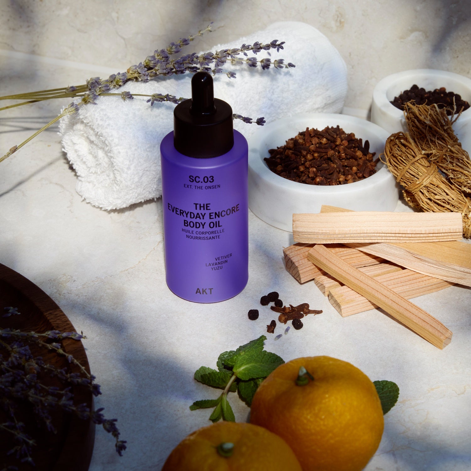 Bottle of AKT The Everyday Encore Body Oil Sc.03 with a black dropper cap stands on a light surface surrounded by spa props: lavender sprigs, white towels, bowls of loose botanicals, wooden sticks, mint leaves, and two oranges. The bottle label reads 'SC.
