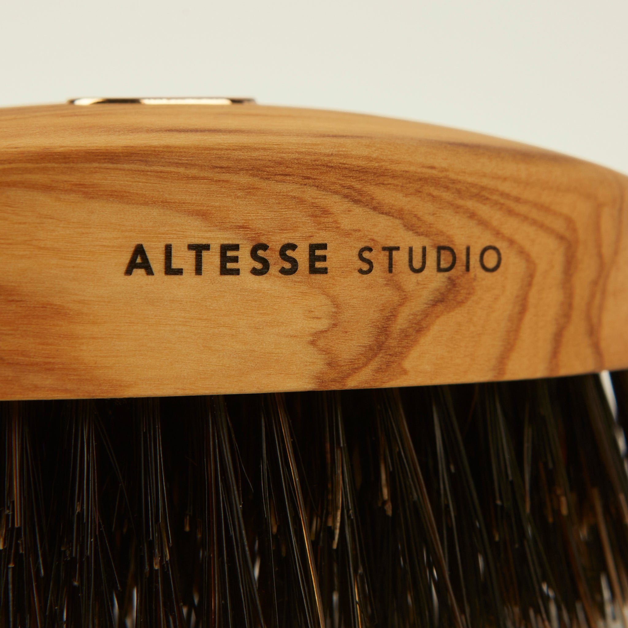 Close-up of a hairbrush with a curved natural wood handle engraved with 'ALTESSE STUDIO' and dark bristles below, on a light neutral background.