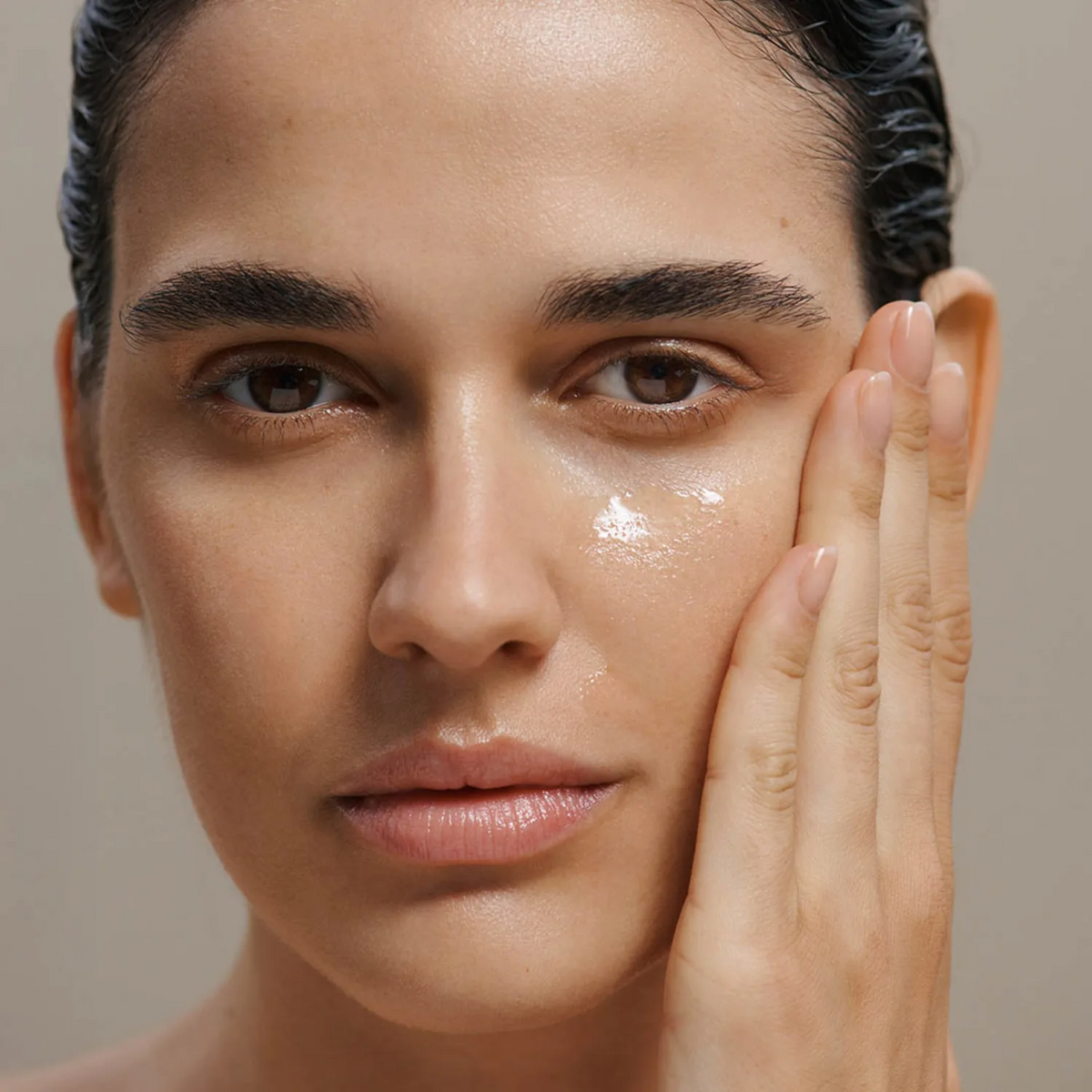 Close-up of a woman's face with a glossy, dewy skincare finish on her cheek, touching her face with her hand against a soft beige background.