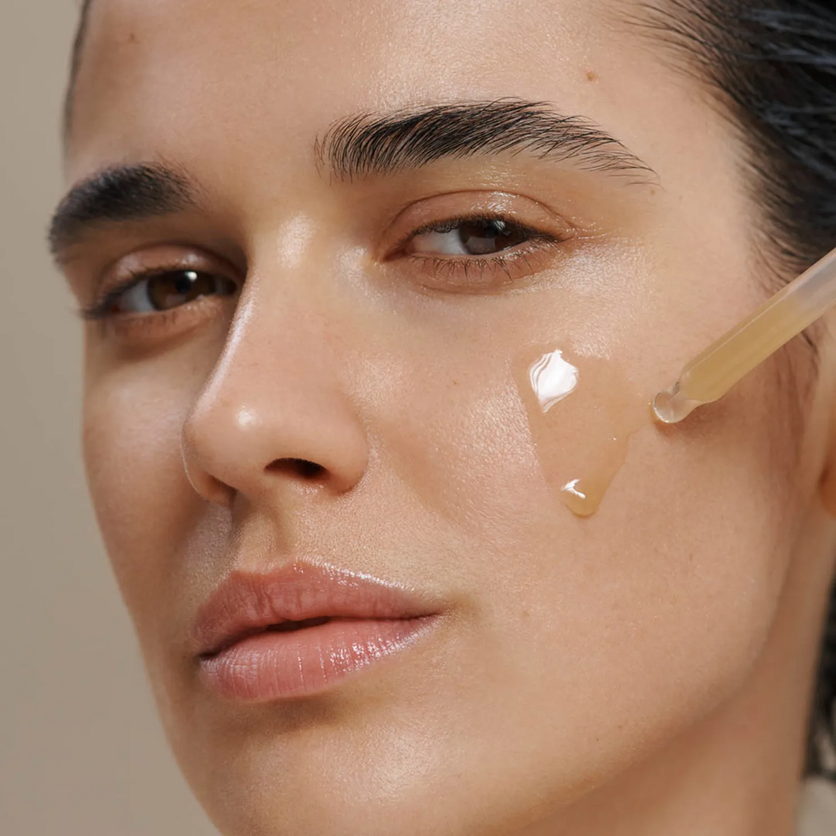 Close-up of a woman's face with a dropper applying a clear gel or serum to her cheek against a soft beige background.
