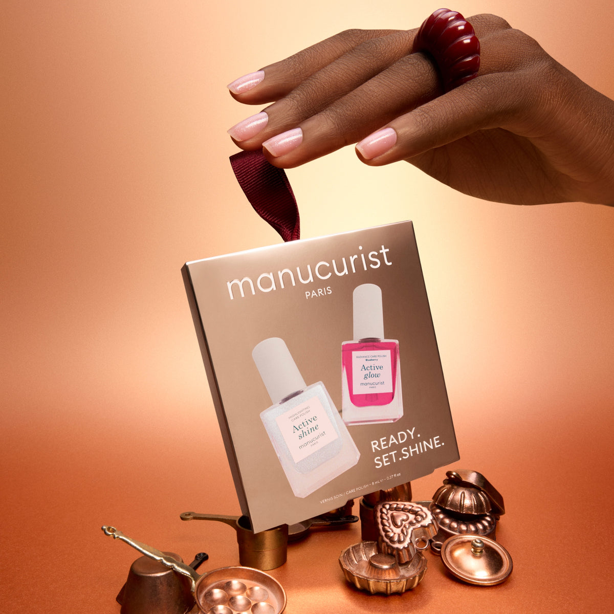 A hand with pale pink manicured nails holds a Manucurist Paris box above a styled arrangement of metallic rose-gold tools and a dark red ring, against a warm peach gradient background.