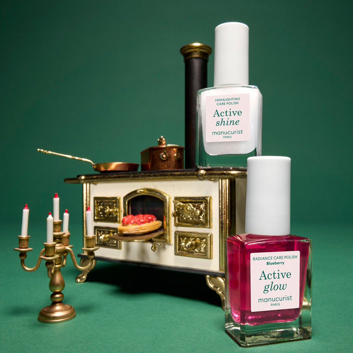 Two manicurist nail polishes by manucurist Paris on a green background: a square bottle of berry pink polish in the foreground and a tall clear bottle of white polish behind it. A miniature stove, candle candelabra, and small decorative objects are arranged around them as styled props.