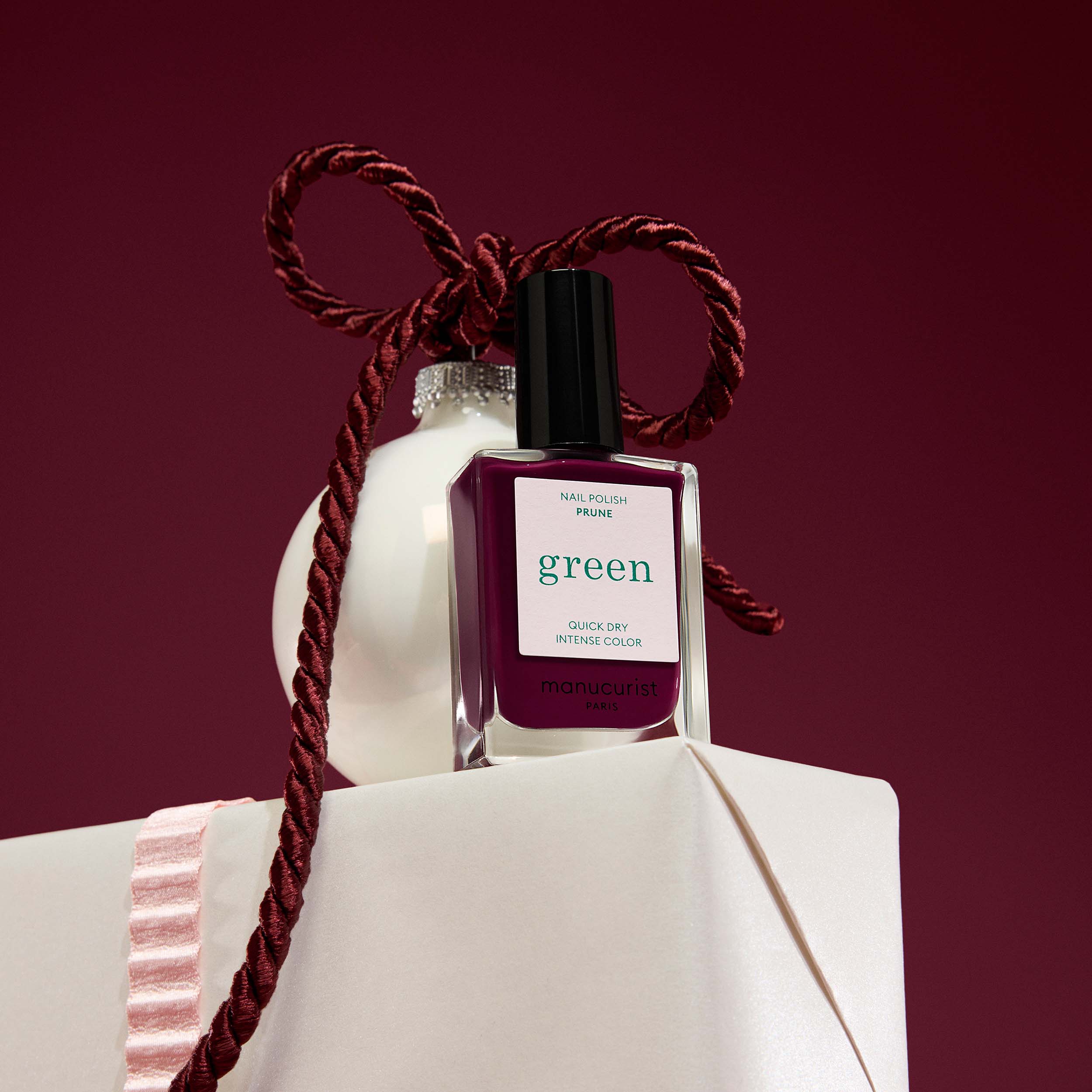 Bottle of Manucurist Prune nail polish with a decorative rope on a white surface against a red background