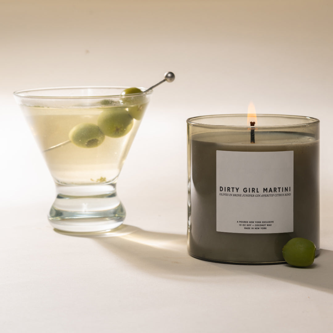 Candle labeled 'Dirty Girl Martini' next to a cocktail with olives on a white background