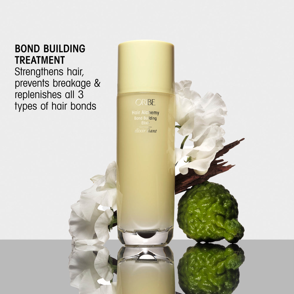 A bottle of Oribe Hair Activator Bond Building Emulsion shown on a reflective surface with white flowers and a green textured prop against a light gray background. Text on the left reads 'BOND BUILDING TREATMENT' and describes strengthening hair and rebuilding bond types.