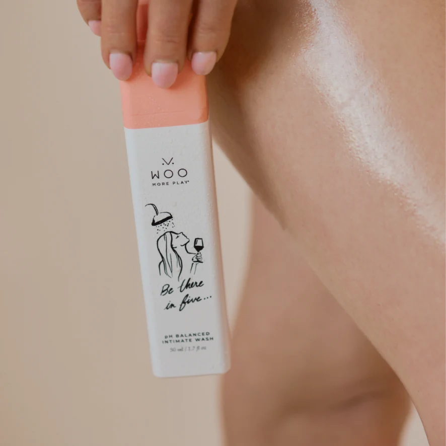 A hand holds a bottle of Woo More Play intimate wash against a close-up of a smooth, glossy leg with a warm beige background.