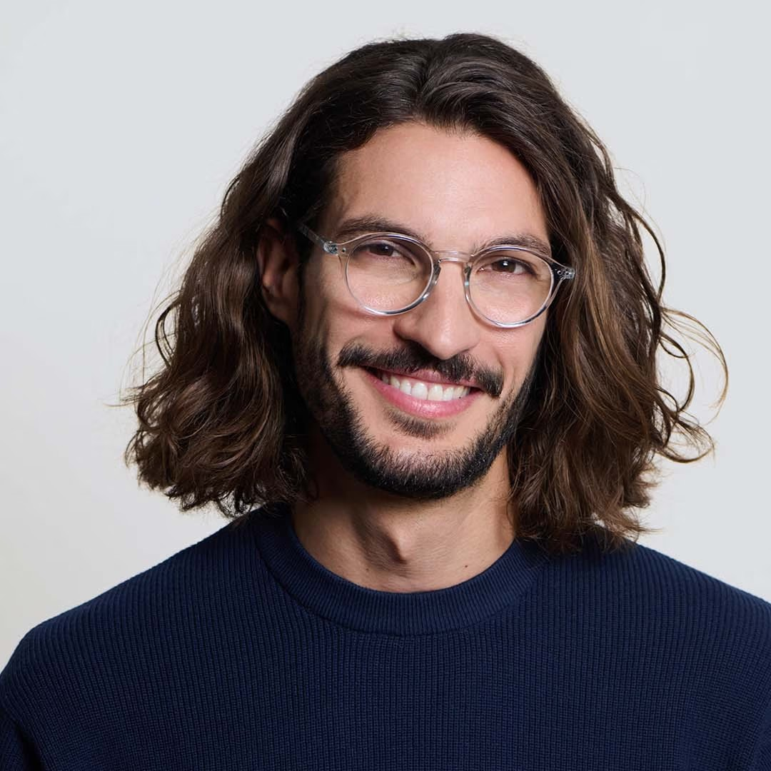 Man with long hair and glasses wearing a navy sweater against a light background