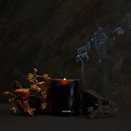 A black glass candle with a small flame, labeled APOTHEKE, styled on a dark surface with dried botanicals and charcoal against a dark textured background with blue smoke rising.