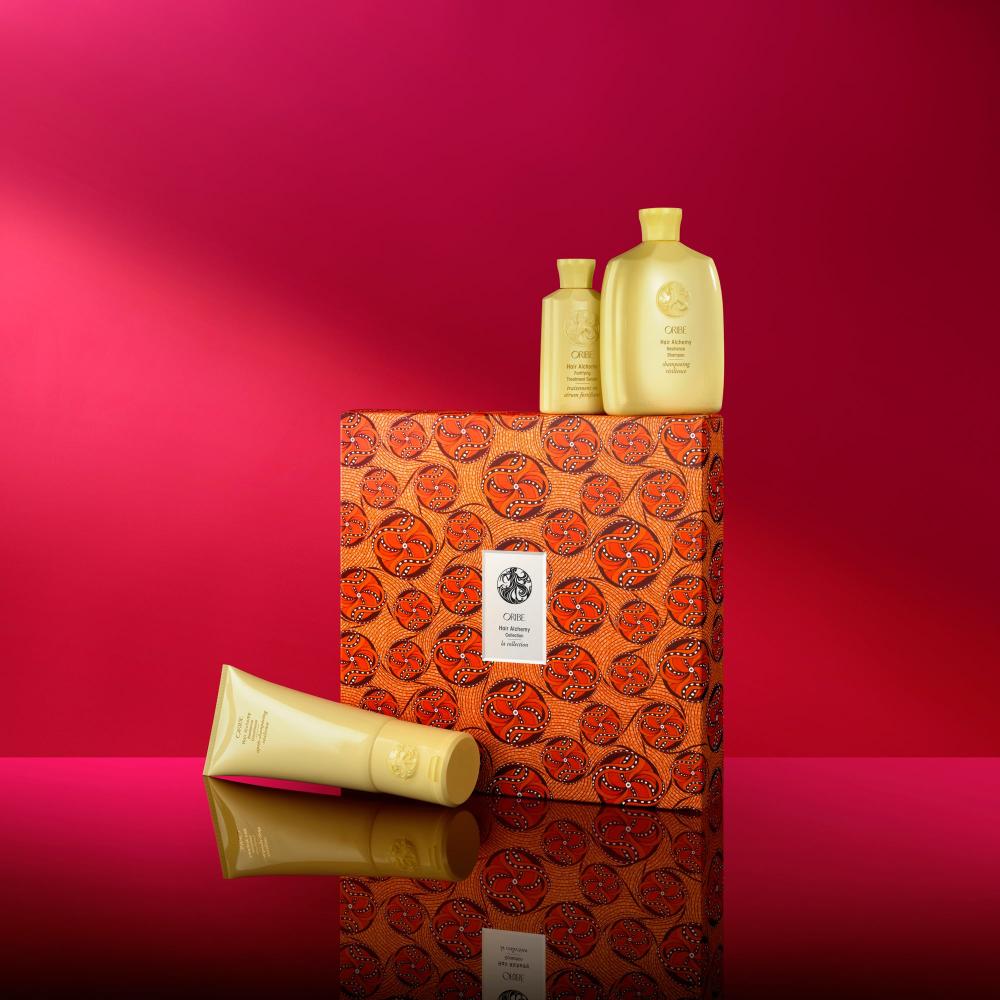 Hair Alchemy Shampoo, conditoner, and serum alongside orange print packaging on red backdrop 