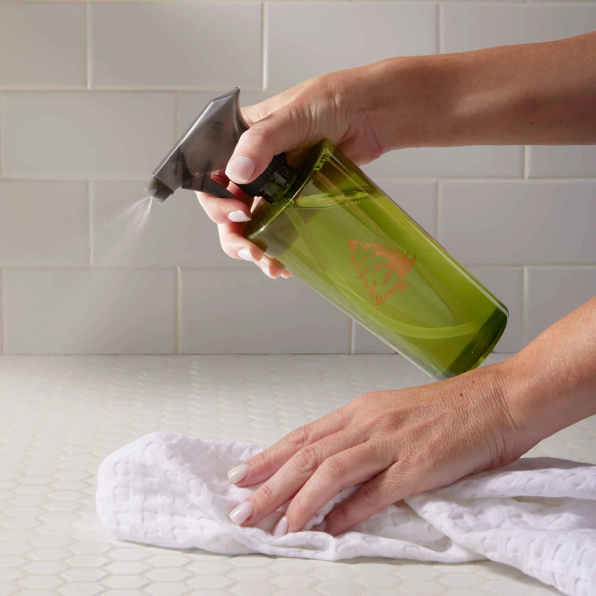 Person cleaning a surface with a green spray bottle and white cloth against a tiled wall background