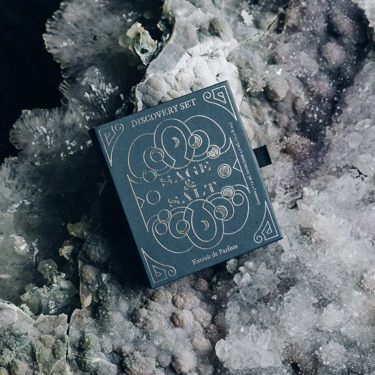A dark teal Discovery Set box by Sage & Salt, featuring intricate silver line art, resting on a textured geode crystal cluster.