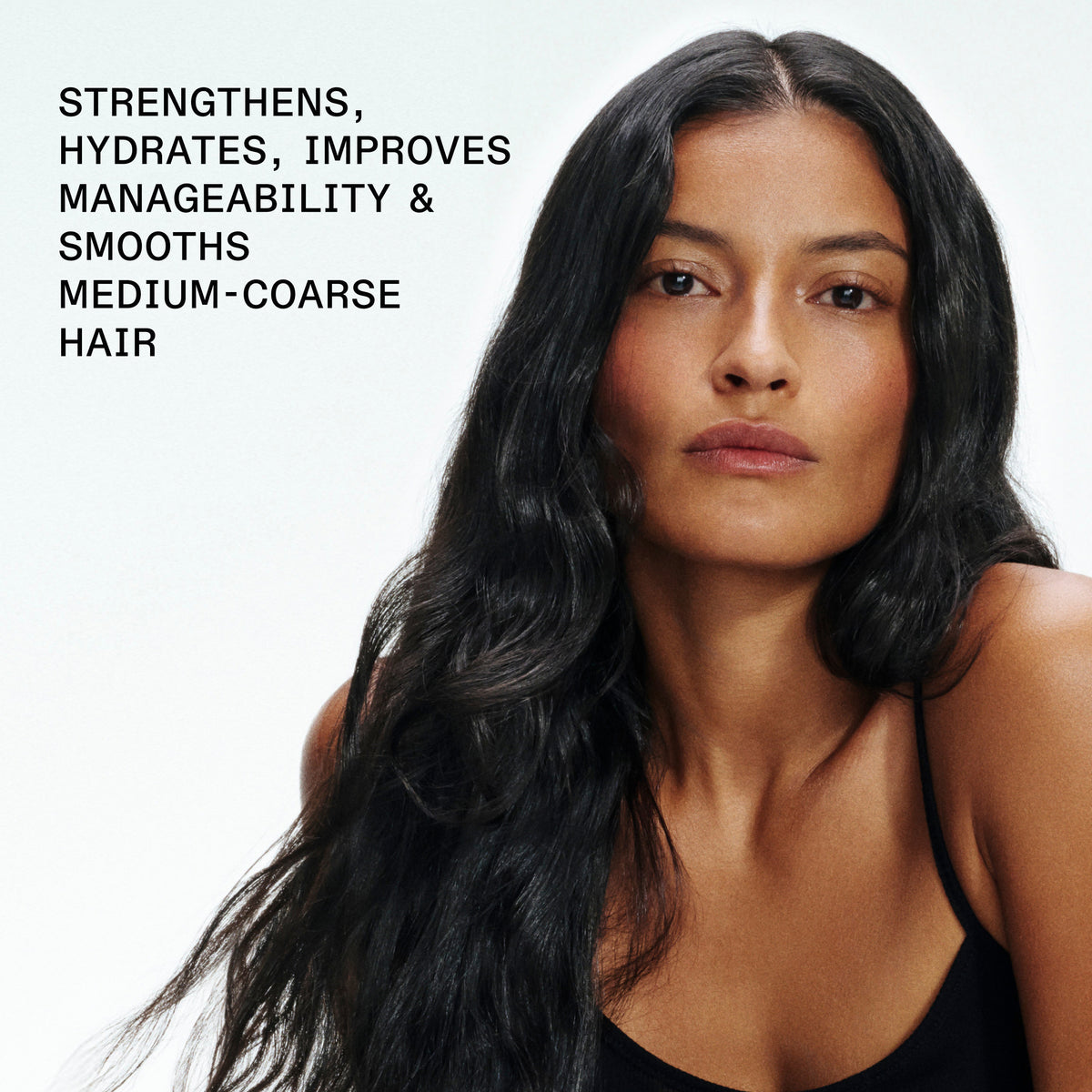 A woman with long, dark, wavy hair wearing a black top, shown against a light background with text on the left that reads: 'STRENGTHENS, HYDRATES, IMPROVES MANAGEABILITY & SMOOTHS MEDIUM-COARSE HAIR'.