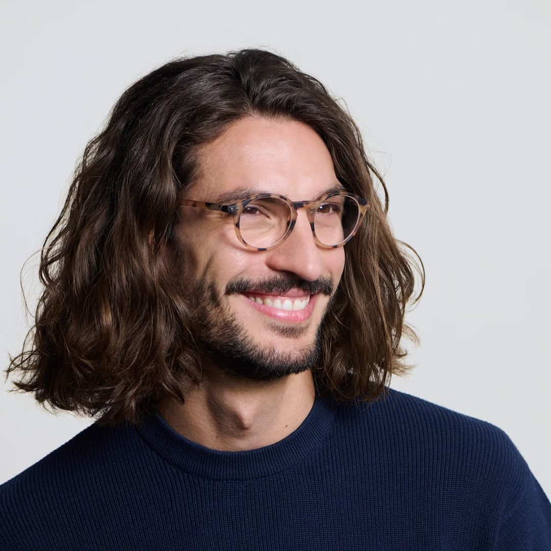 Man with long hair and glasses wearing a navy sweater against a plain background