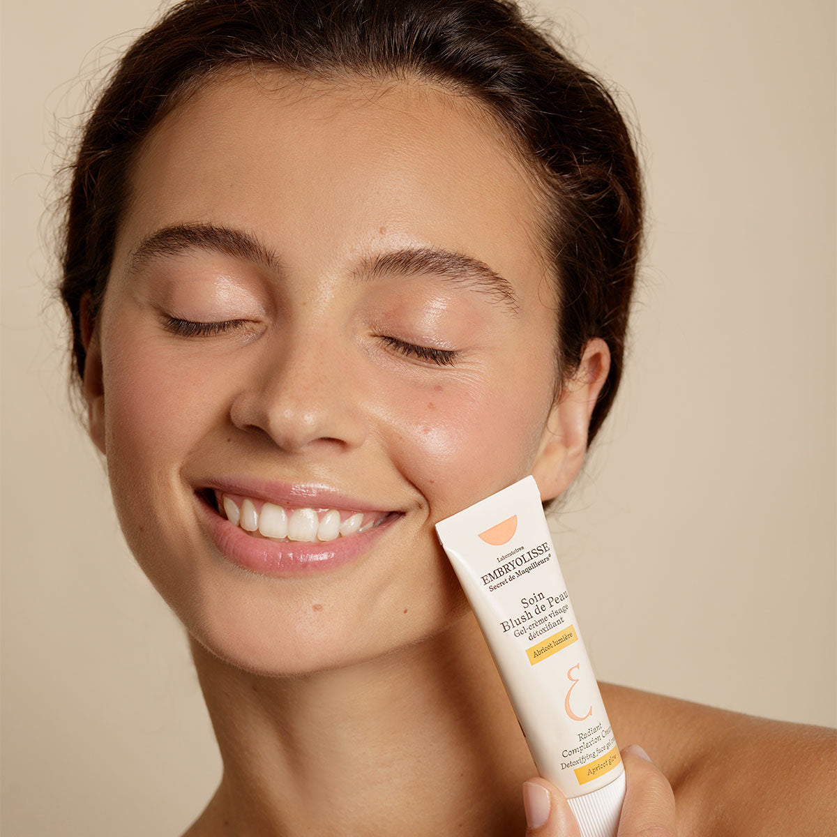 Woman smiling and holding up a bottle of Embryolisse Radiant Complexion Cream in Apricot Glow.