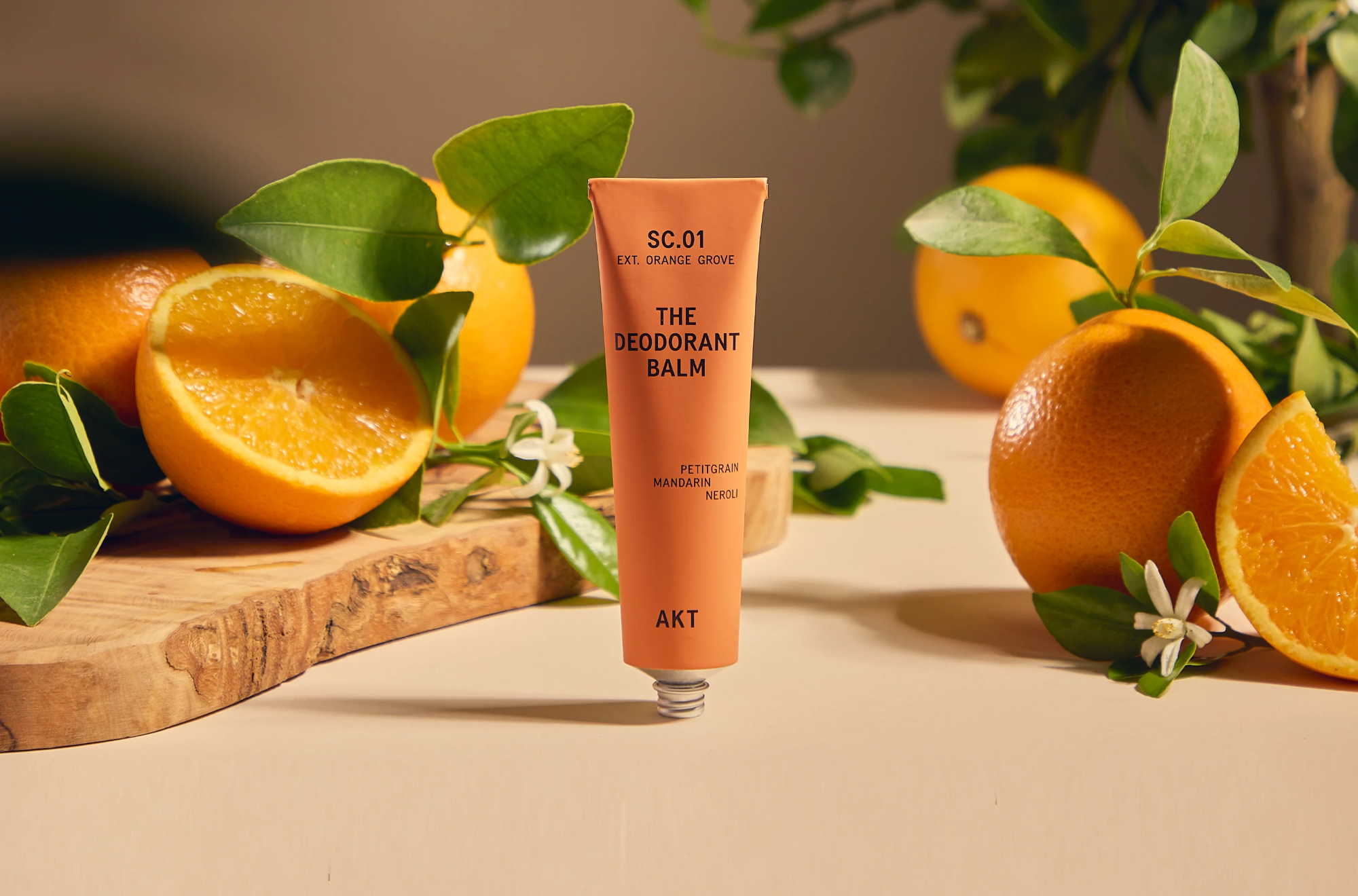 AKT Scene 1 Orange Grove Deodorant Balm on tan surface in front of fresh oranges.