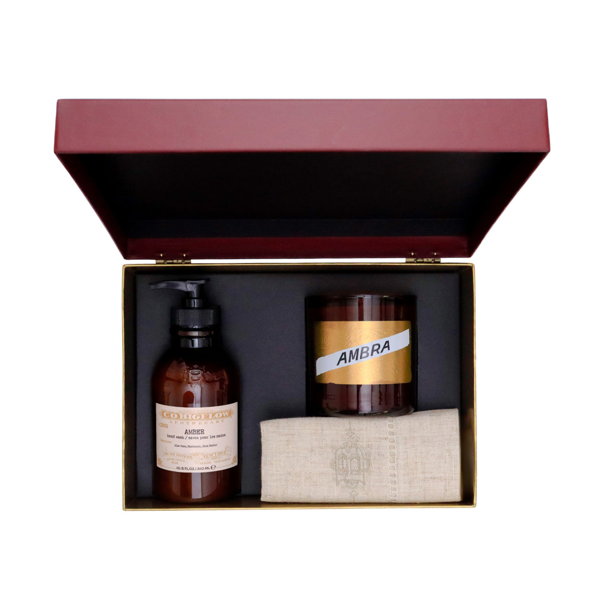 A luxury gift set in a dark red and gold presentation box, containing an amber glass bottle of CO Bigelow Handmade soap, an amber glass candle labeled Ambra, and a beige linen cloth.