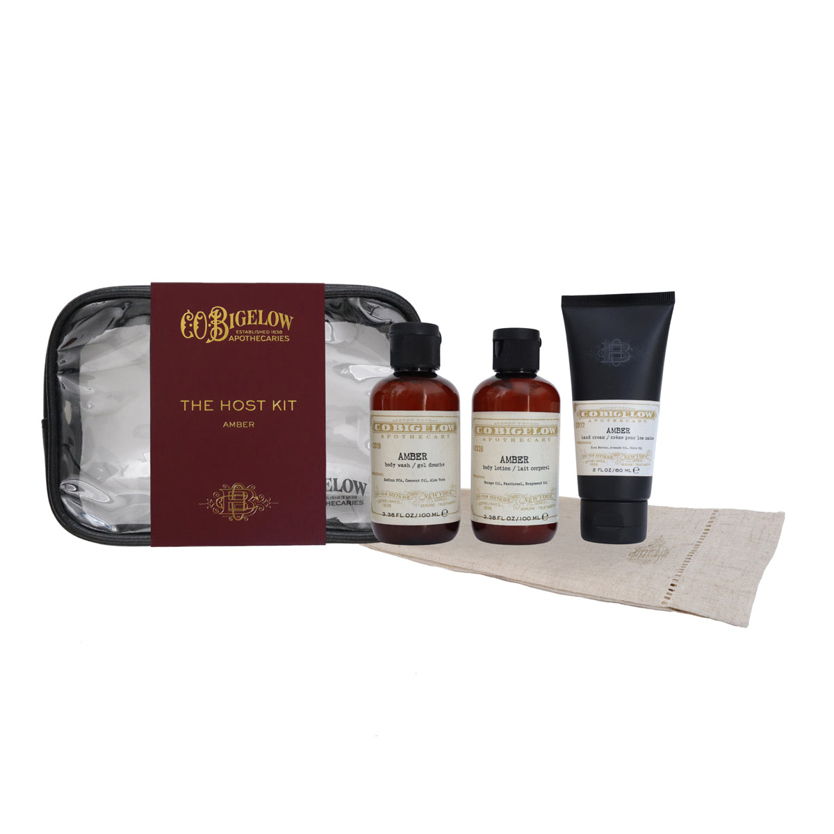 Bigelow Apothecary 'The Host Kit' amber set in Amber, featuring two amber bottles of shower liquid, a black tube of body wash, a black zippered case with a burgundy label, and a beige towel on a white background.