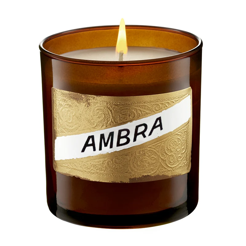 An amber glass jar candle with a single lit wick, featuring a decorative gold foil label wrapped around the middle with a white diagonal banner that reads 'AMBRA' in black text, set against a plain white background.