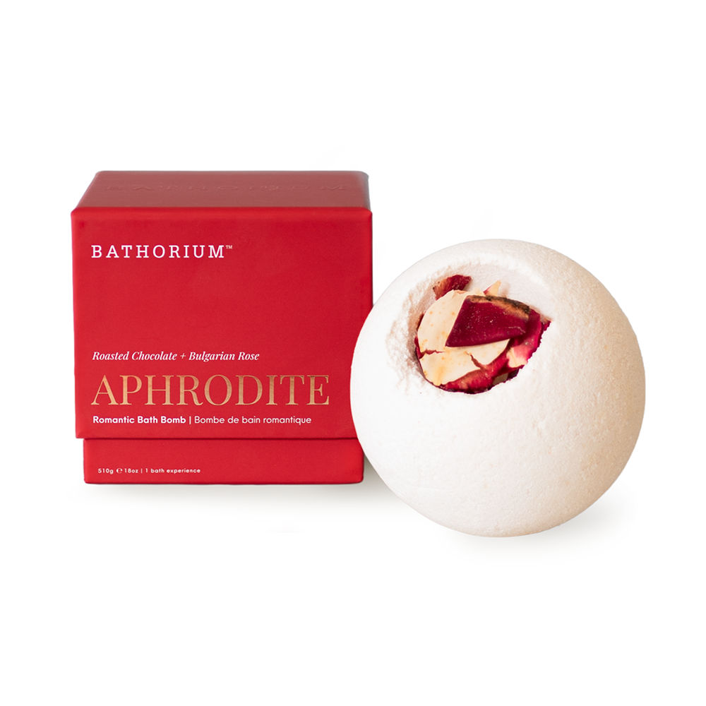 A Bathorium Aphrodite romantic bath bomb next to its red packaging box on a white background. The spherical bath bomb is off-white with a cut opening showing red and beige fruit pieces inside, and the box is bright red with gold lettering.