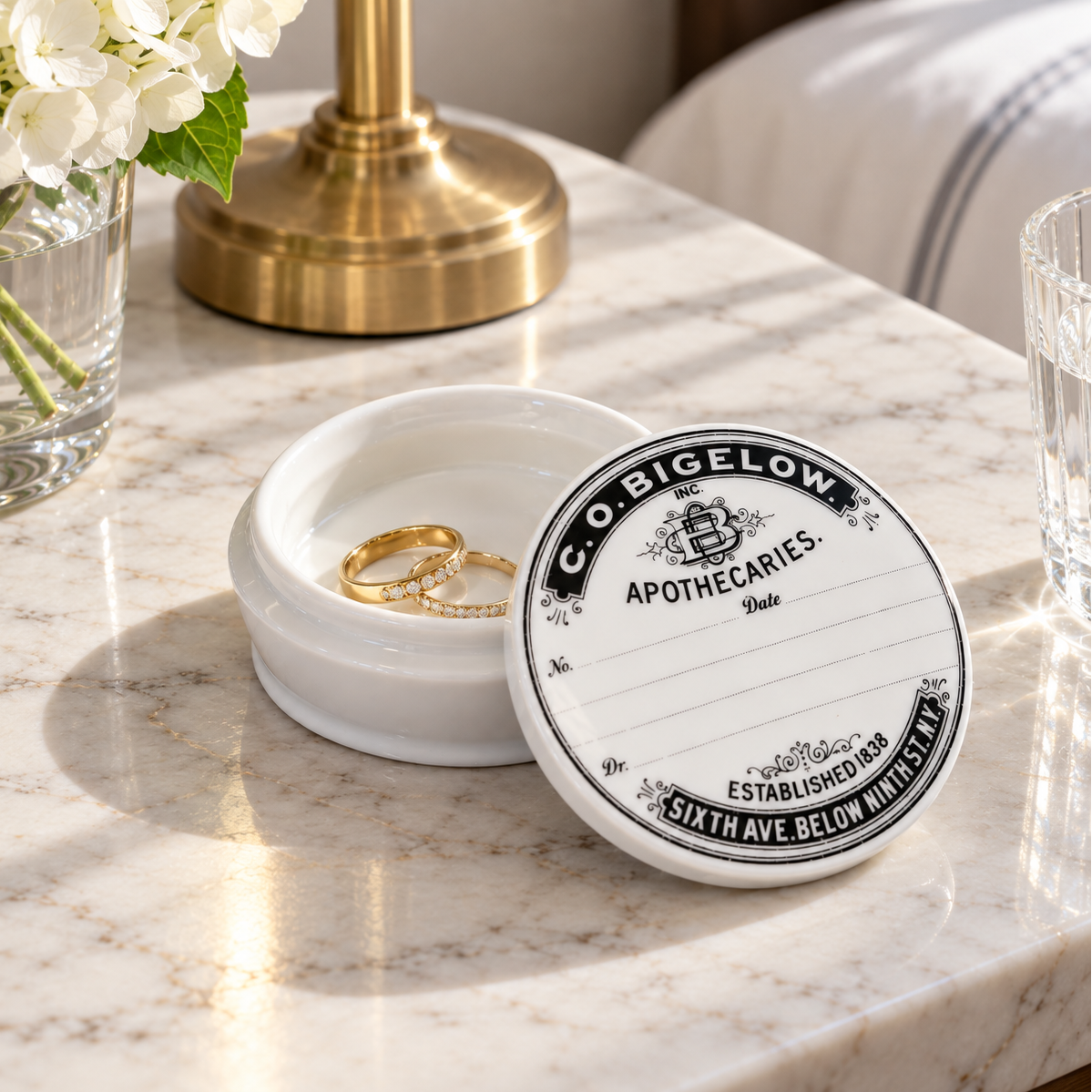 A white ceramic ring box with a lid printed with vintage-style apothecary text, shown on a marble surface beside a glass vase of white flowers and a brass lamp base. Two gold rings are placed inside the box.