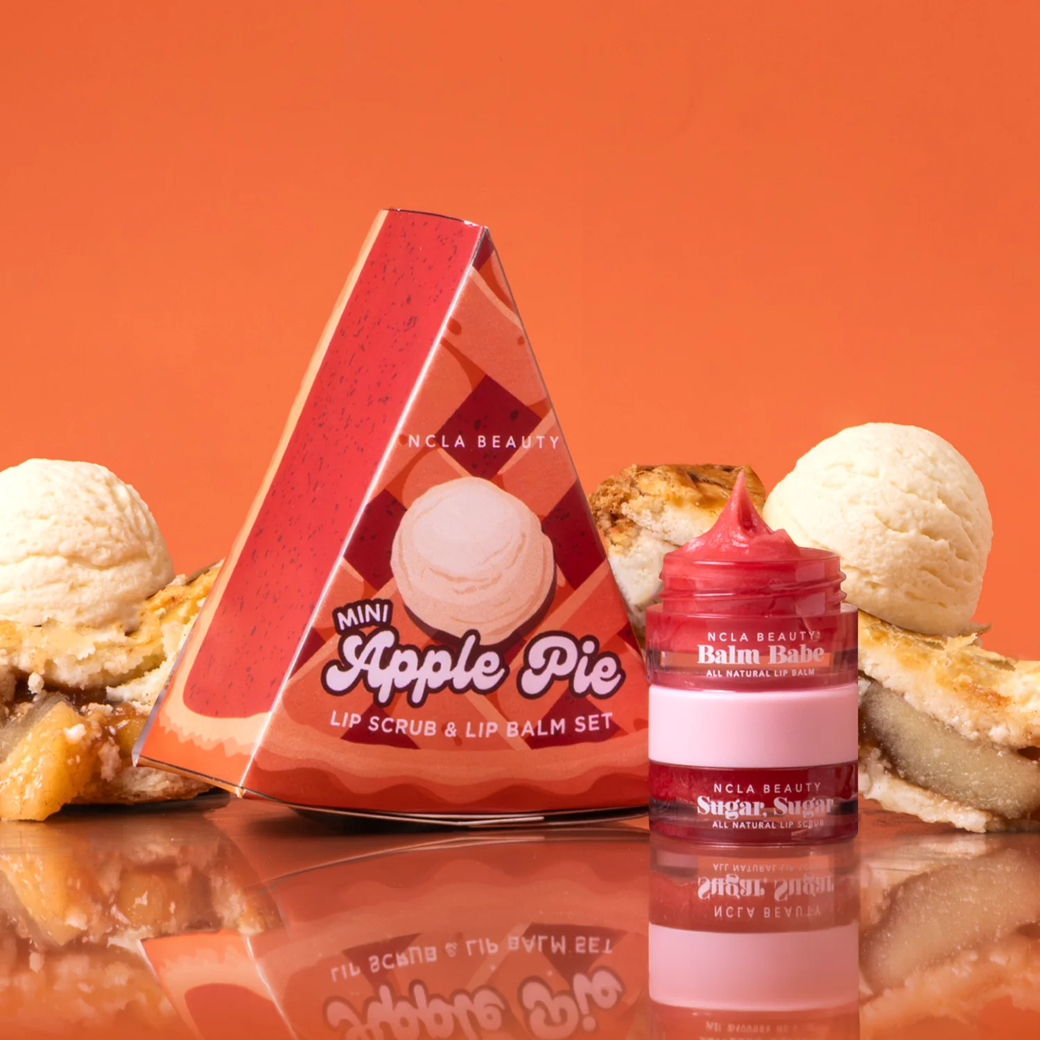 NCLA Beauty Mini Apple Pie lip scrub and lip balm set, featuring a triangular red and orange box with an apple pie illustration next to an open jar of red balm and a pink lip scrub.