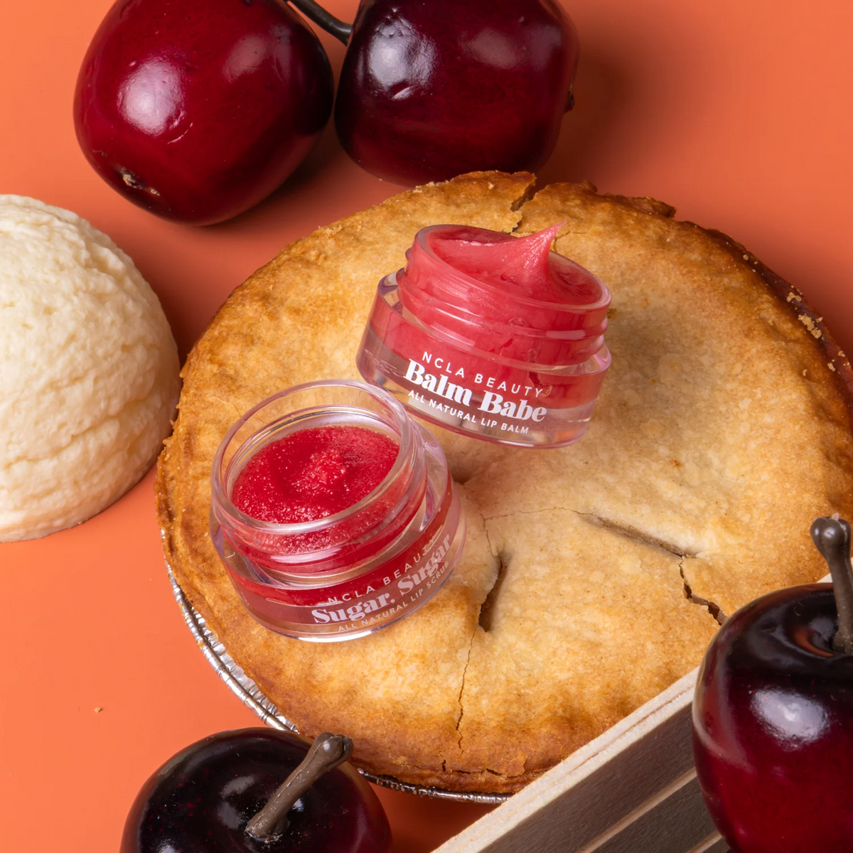 Two small jars of NCLA  Beauty 'Balm Babe' sugar mask balm, one with a pinkish-red balm and one with a bright red glittery balm, styled on a golden-brown pie crust with whole cherries and a scoop of vanilla ice cream on an orange background.