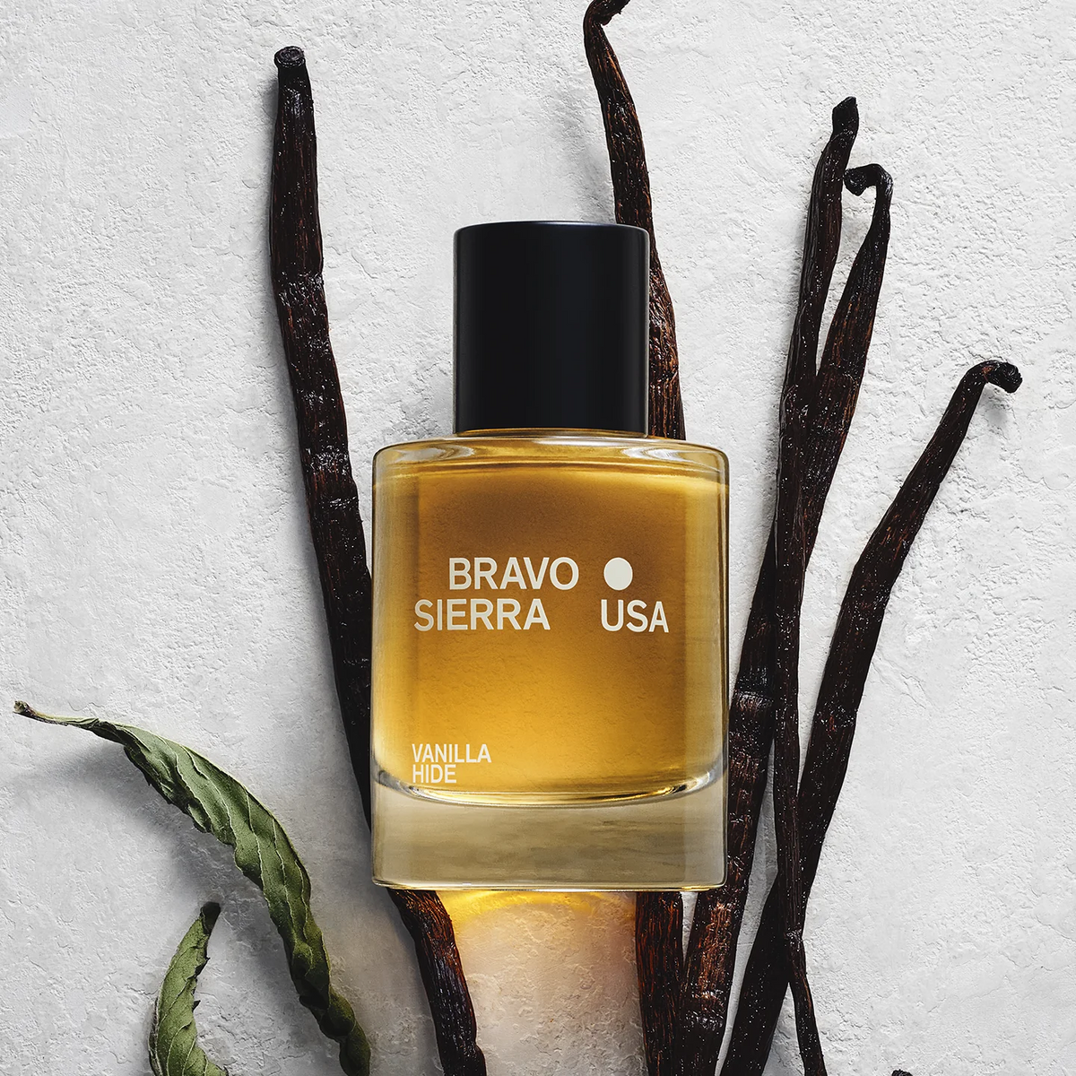 A bottle of Bravo Sierra Vanilla Hide - Eau de Parfum with amber liquid and a black cap, styled on a textured white background with vanilla beans.