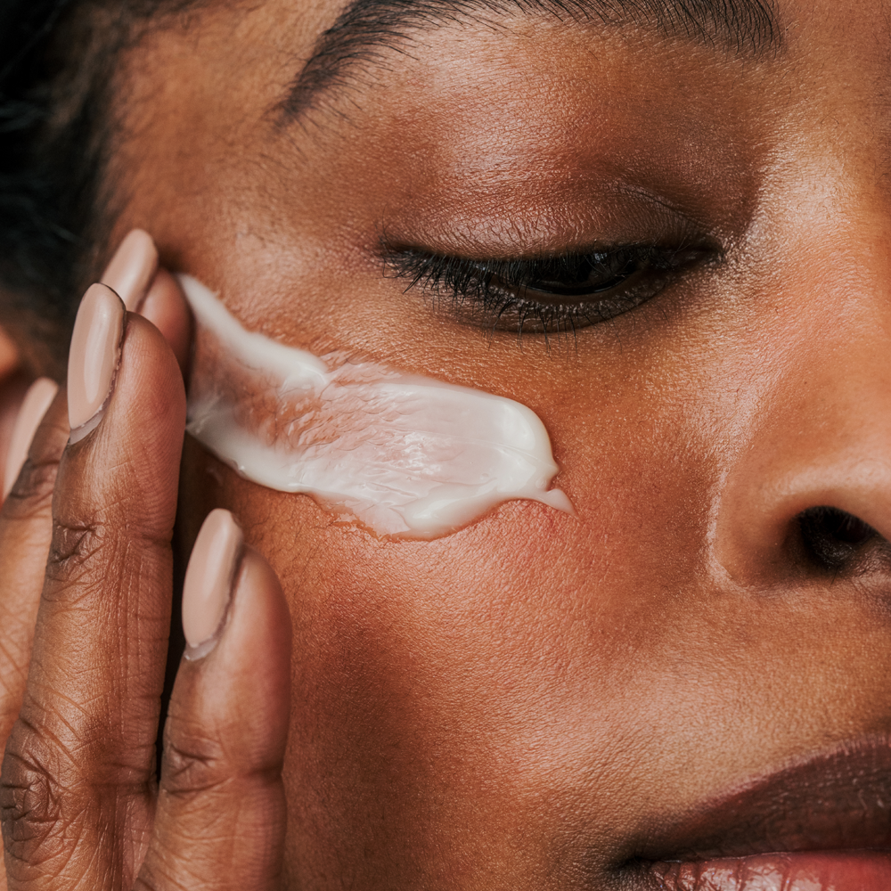 Close-up of a person applying or spreading Radford Butter -  The Thick Cream across their cheek with a hand, showing skin texture and makeup details.