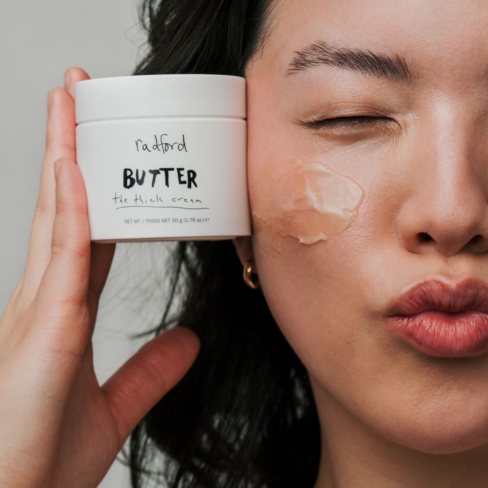 A close-up lifestyle shot of a woman applying or wearing a clear cream patch on her cheek, holding a white jar of Radford Butter -  The Thick Cream. The background is a soft, neutral gray.