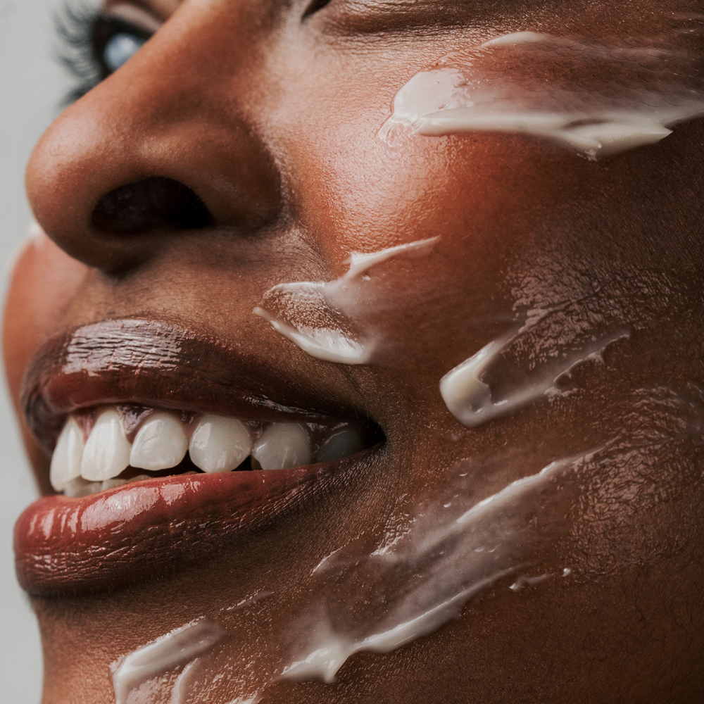 Close-up of a person's face with Radford Butter -  The Thick Cream smeared across the cheek and lips, showing rich skin texture and glossy makeup.