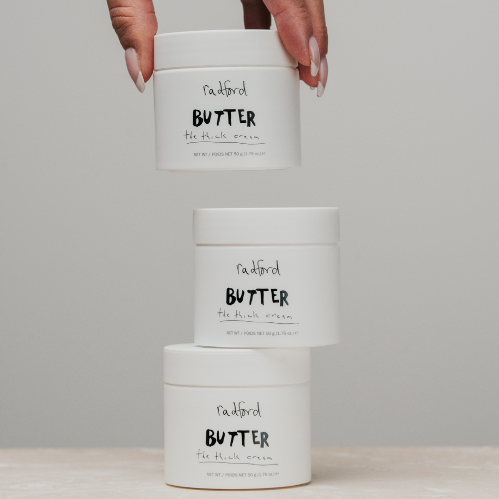 Three white jars of Radford Butter -  The Thick Cream stacked in a tower, with a hand holding the top jar against a neutral gray background.